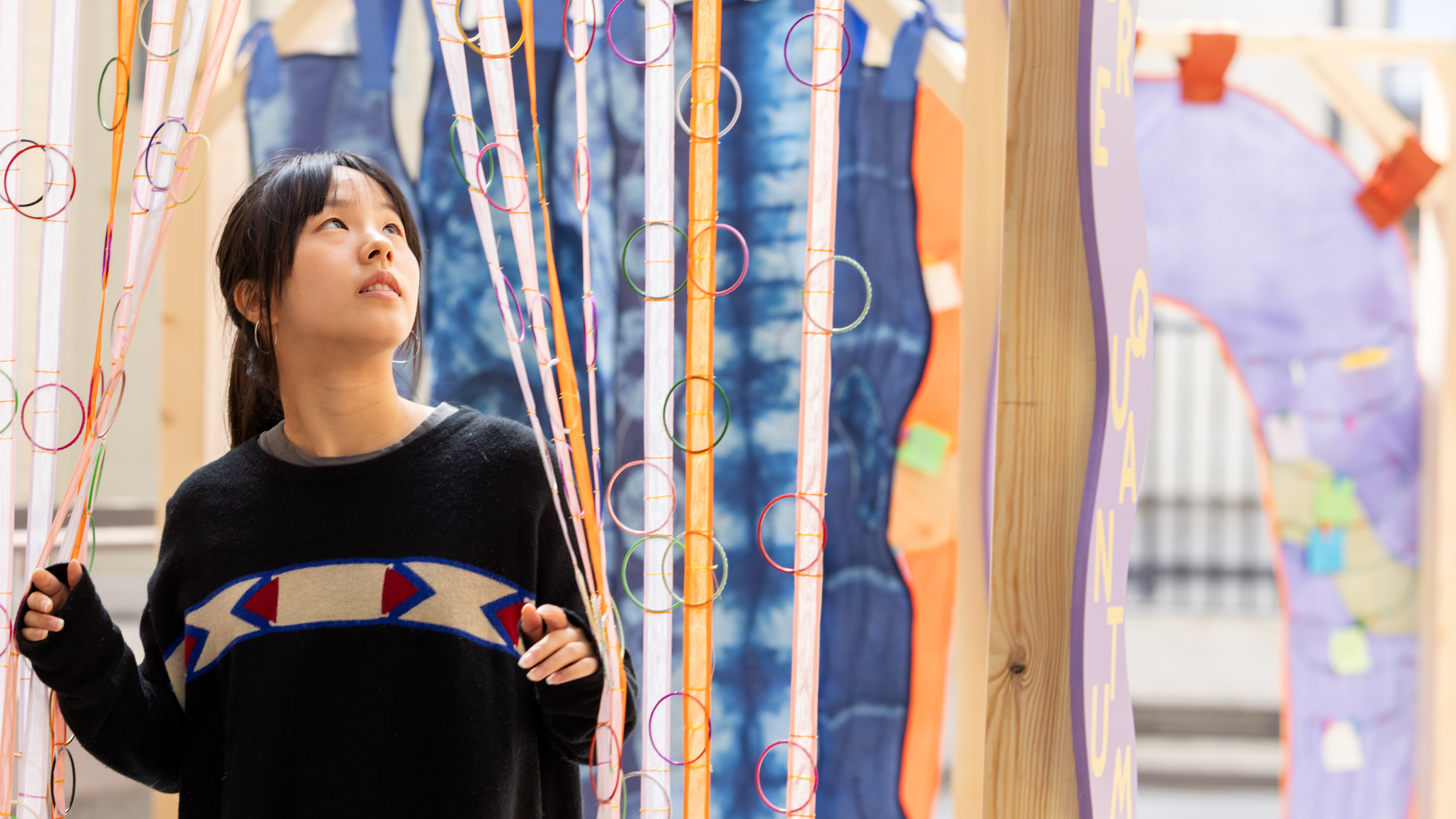 A young adult goes through a curtain of colourful streams of fabric hanging from a gate-like wooden structure.