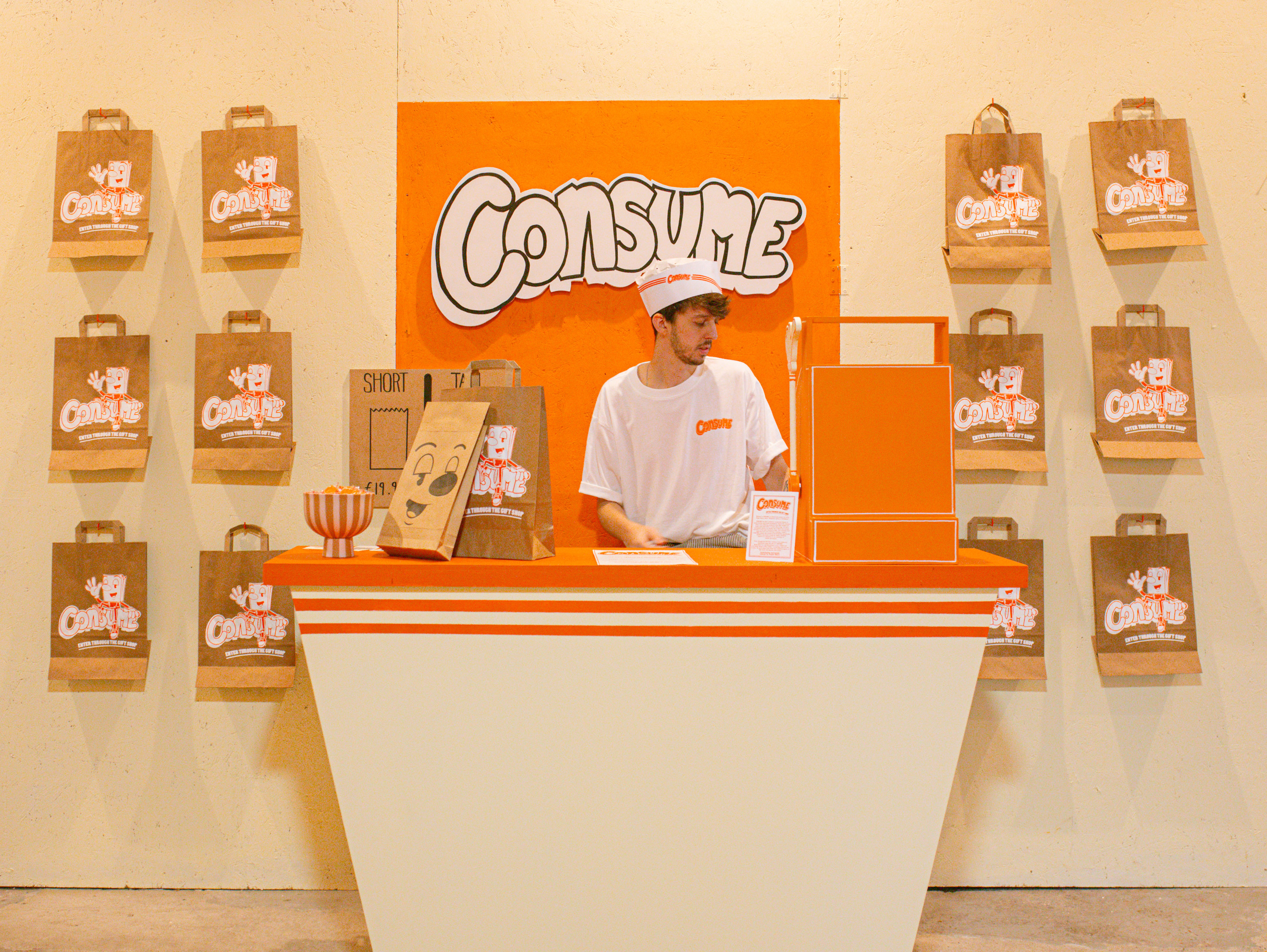 An orange and white counter, a sign "consume" hung behind. printed brown paper bags to either side