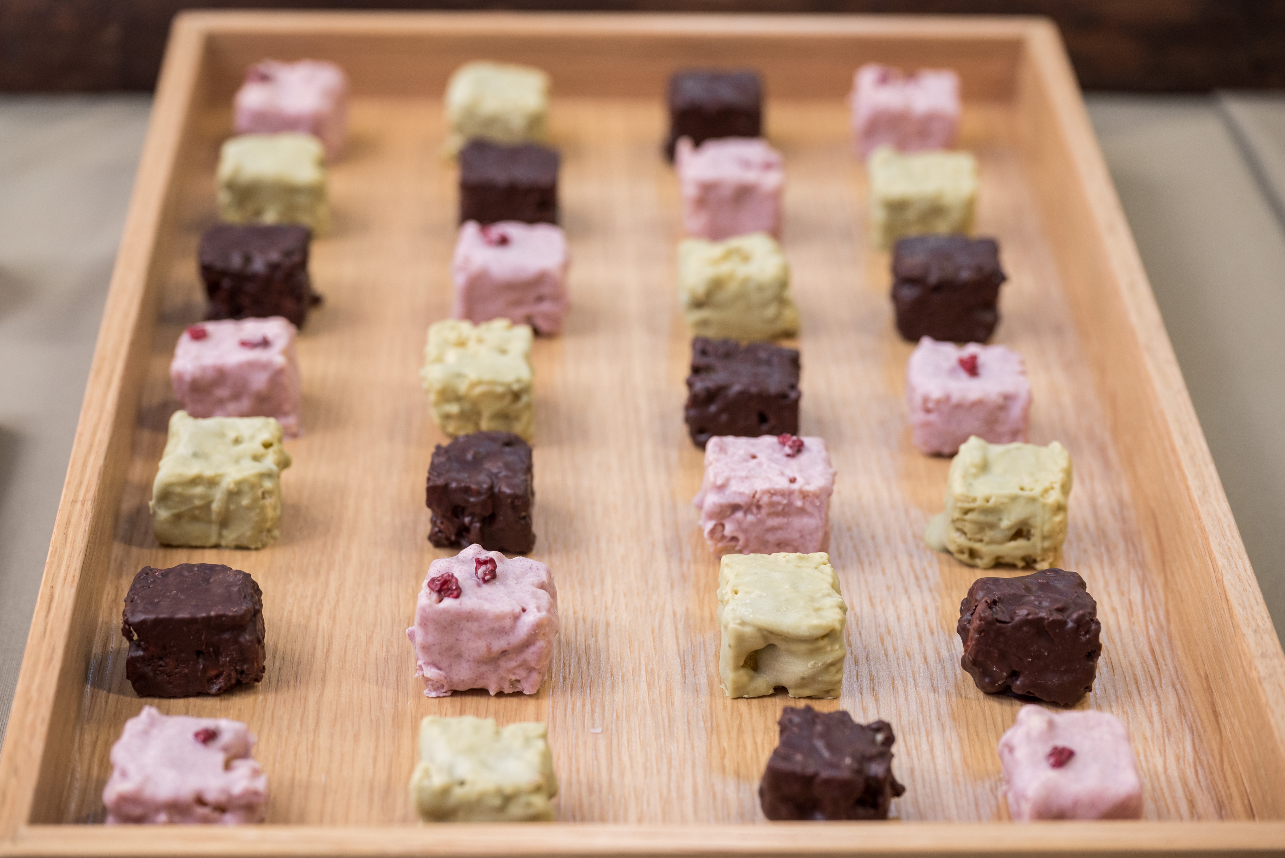 A wooden tray with a grid of bite-sized cakes. The small cubes come in three flavours: dark chocolate, pink, and green.