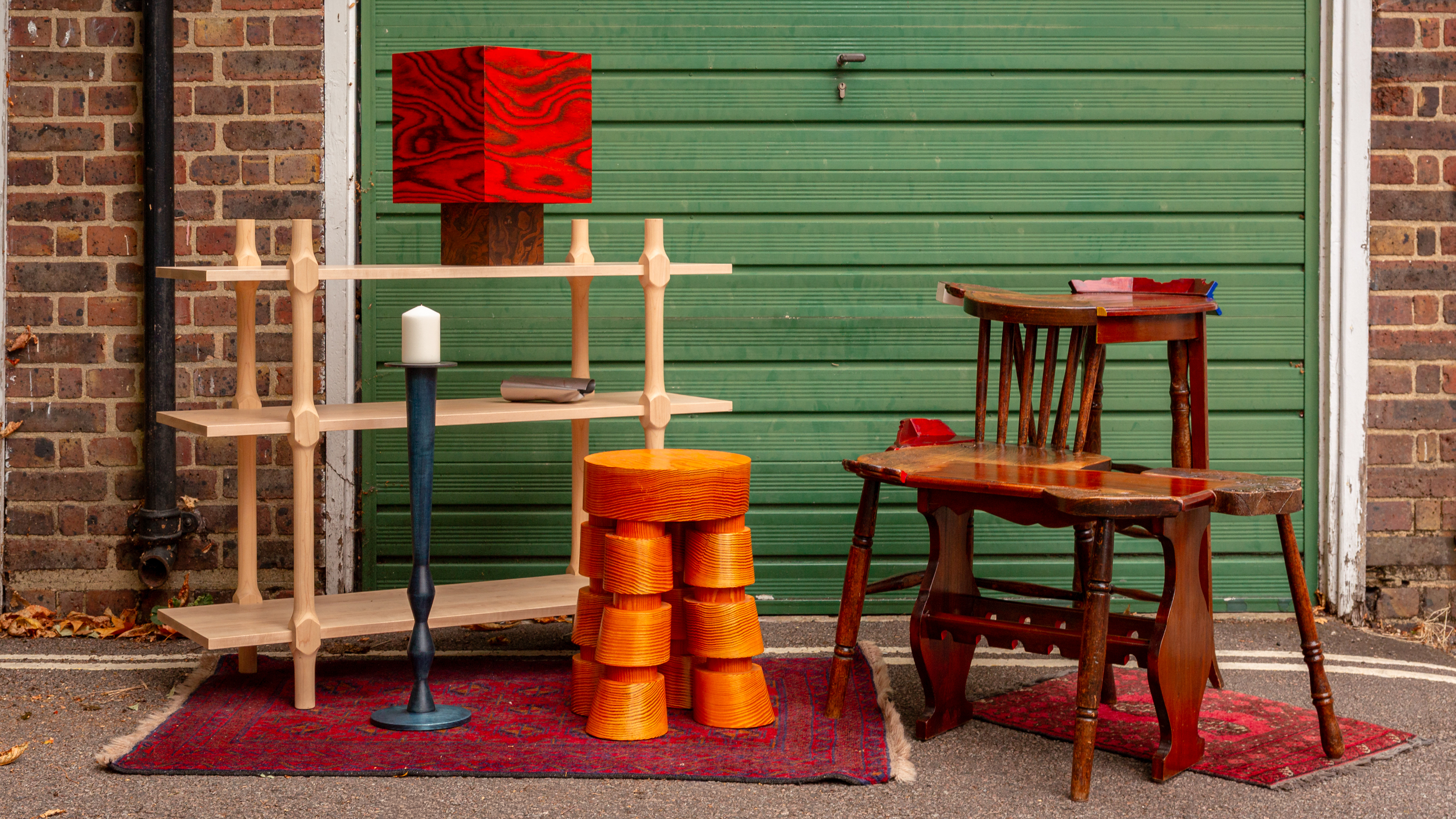 Eclectic furniture on red rugs before a green garage door, featuring wooden shelf, orange stools, and mismatched chair.