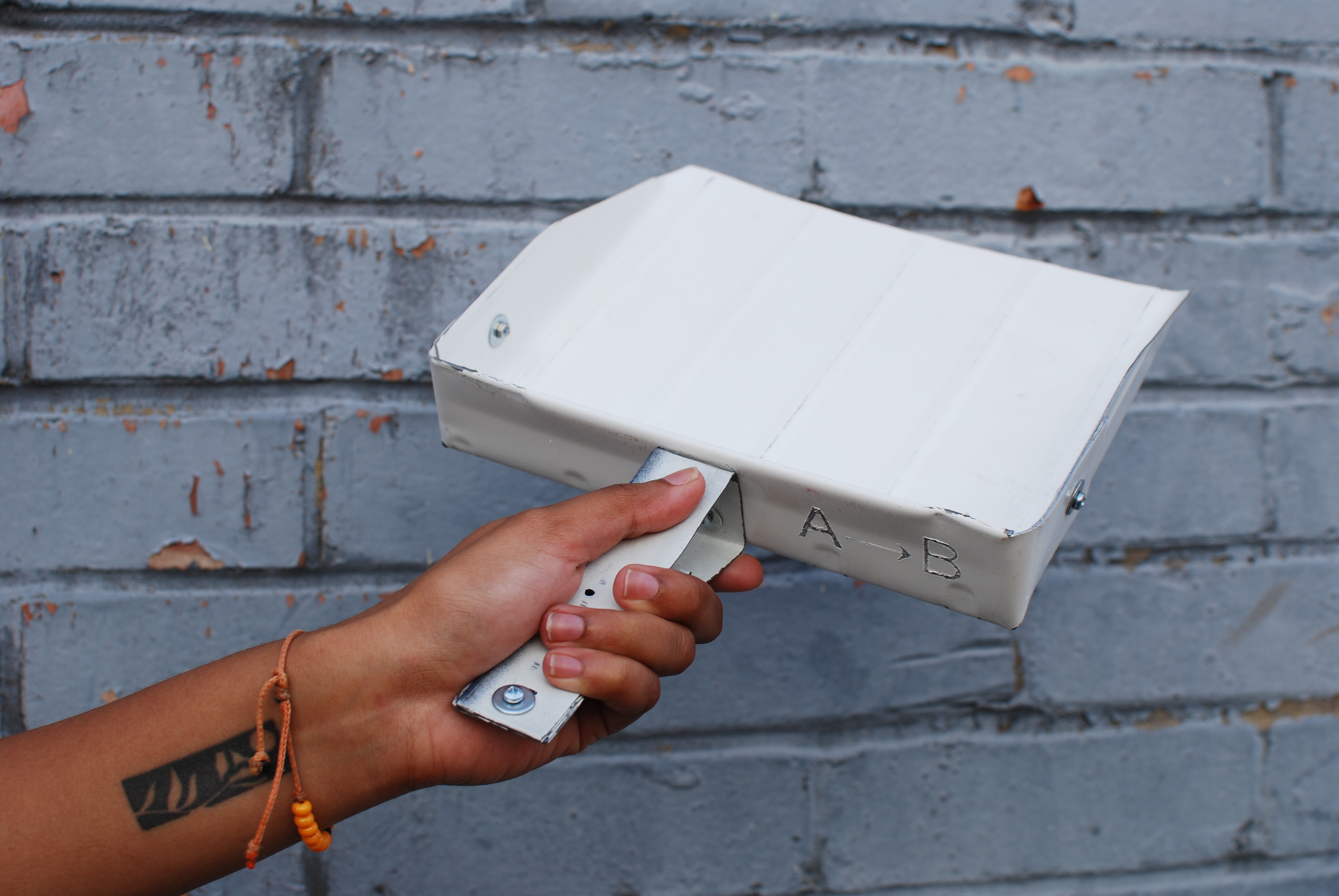 A young person holds up a prototype dustpan, made of reclaimed white metal, in front of a grey brick wall.